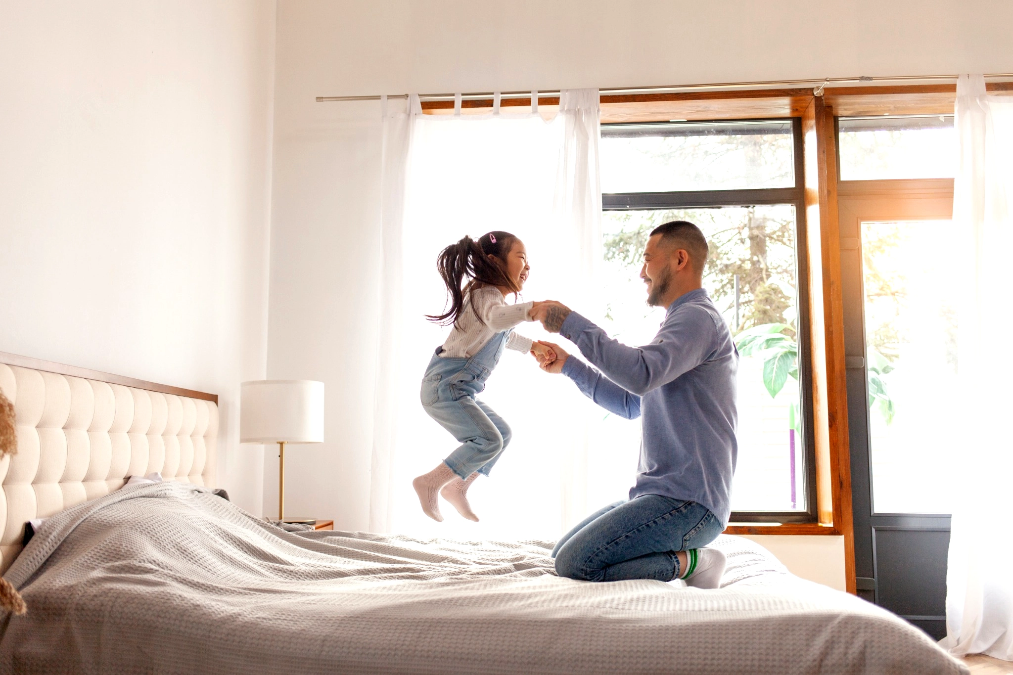 Father holding young daughter's hands while she jumps on the bed