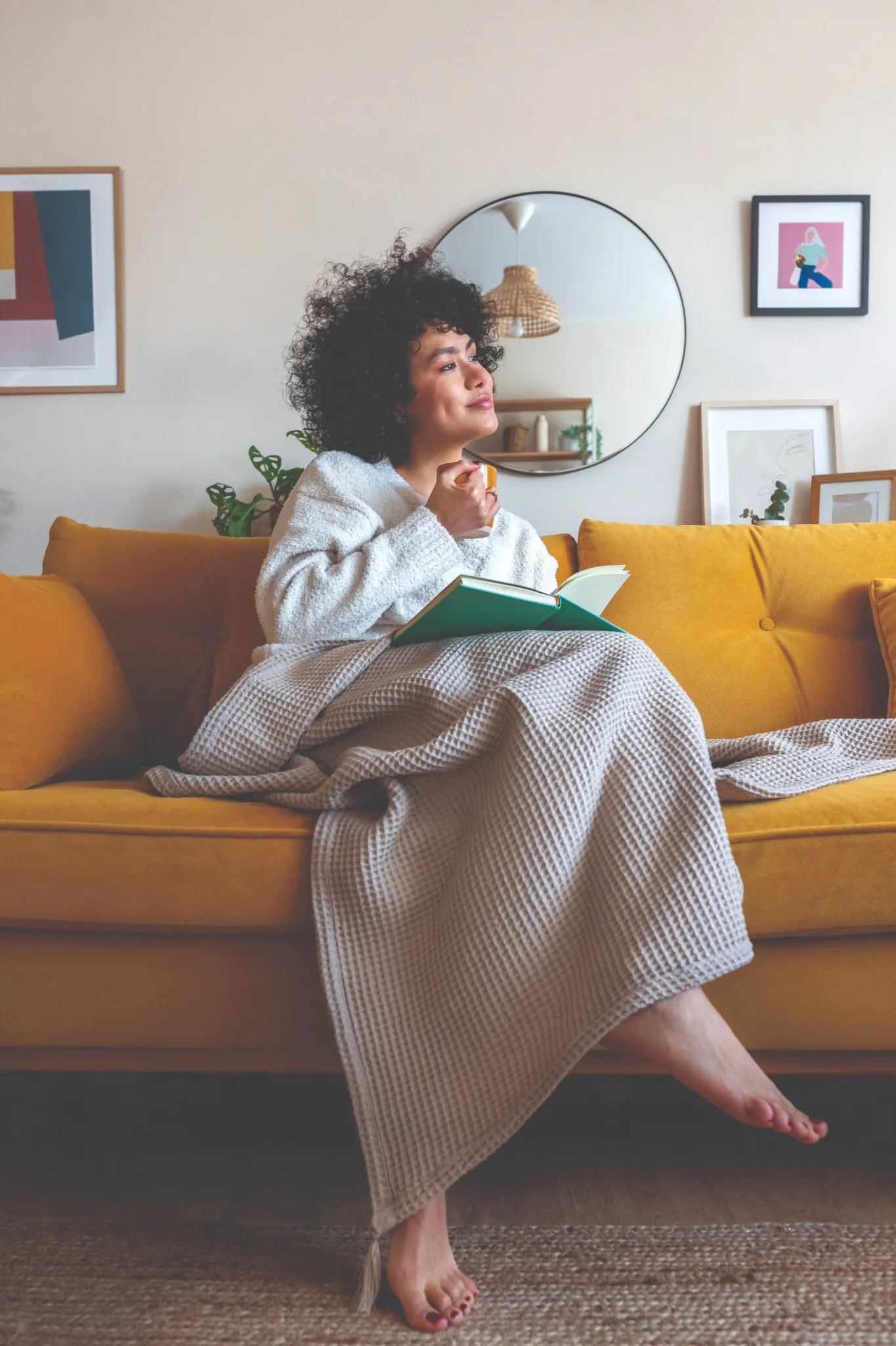 Contented woman snuggled up on yellow couch with a blanket, warm drink, and book