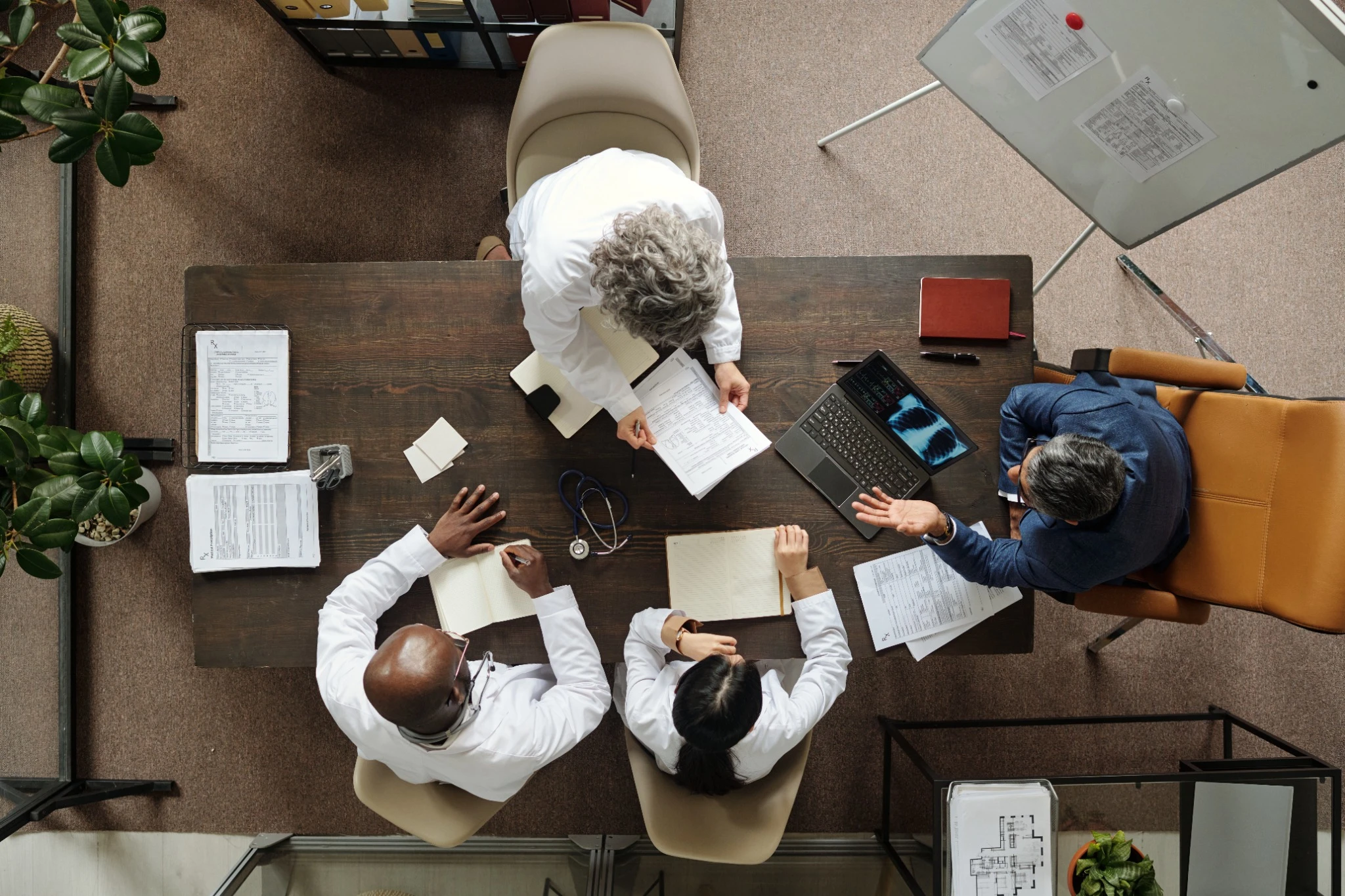 Group of middle aged and senior multiethnic doctors and one middle aged Caucasian man sitting around table discussing medical documents and using laptop during meeting, top view.