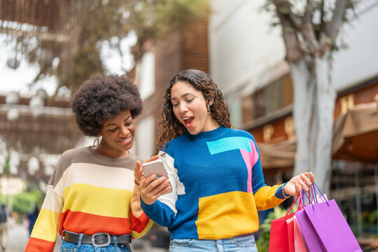 Two young women walking down sidewalk with shopping bags looking at phone