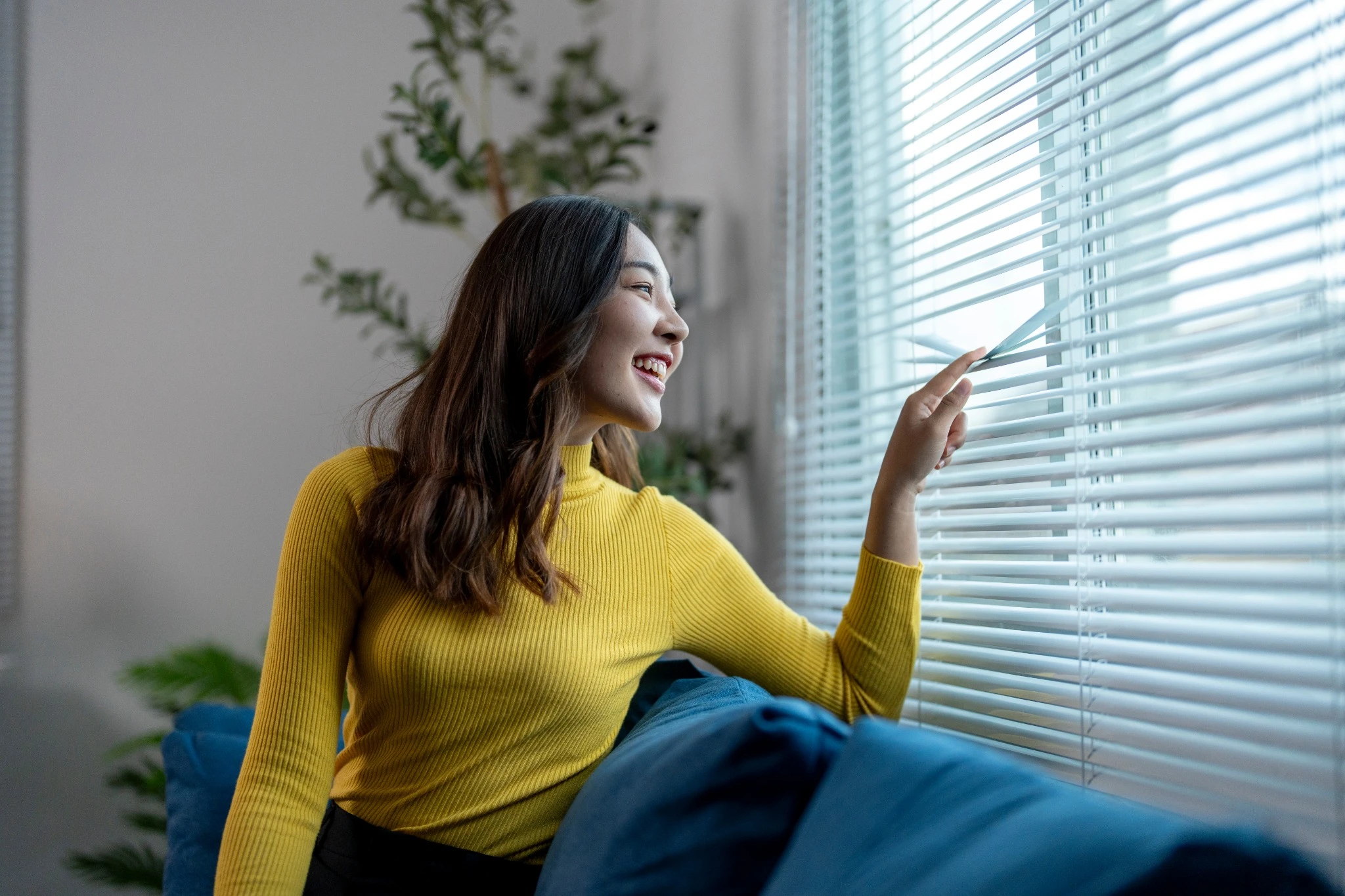 Young woman sitting on sofa is opening blinds and looking through the window.