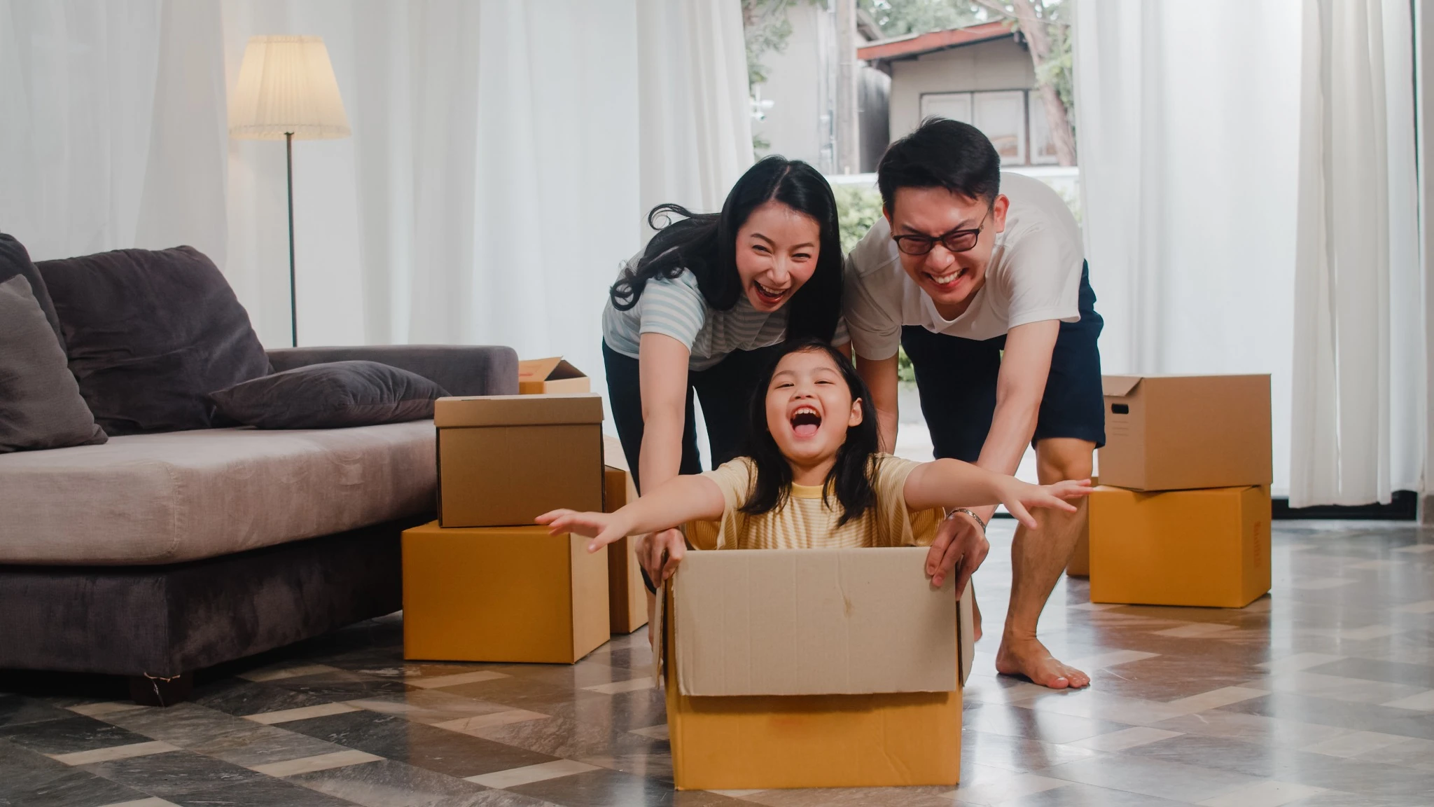 Happy Asian young family having fun laughing moving into new home. Japanese parents mother and father smiling helping excited little girl riding sitting in cardboard box. New property and relocation..