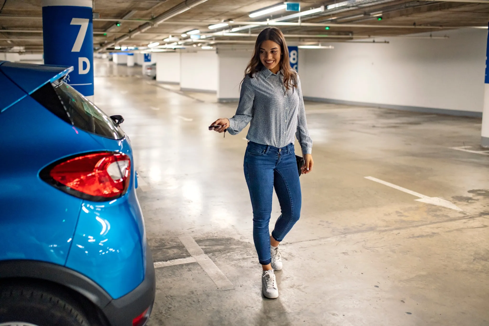 Woman in a parking, unlocking in her car. Woman activating her car alarm in an underground parking garage as she walks away. Young business woman walking with car keys in the underground
