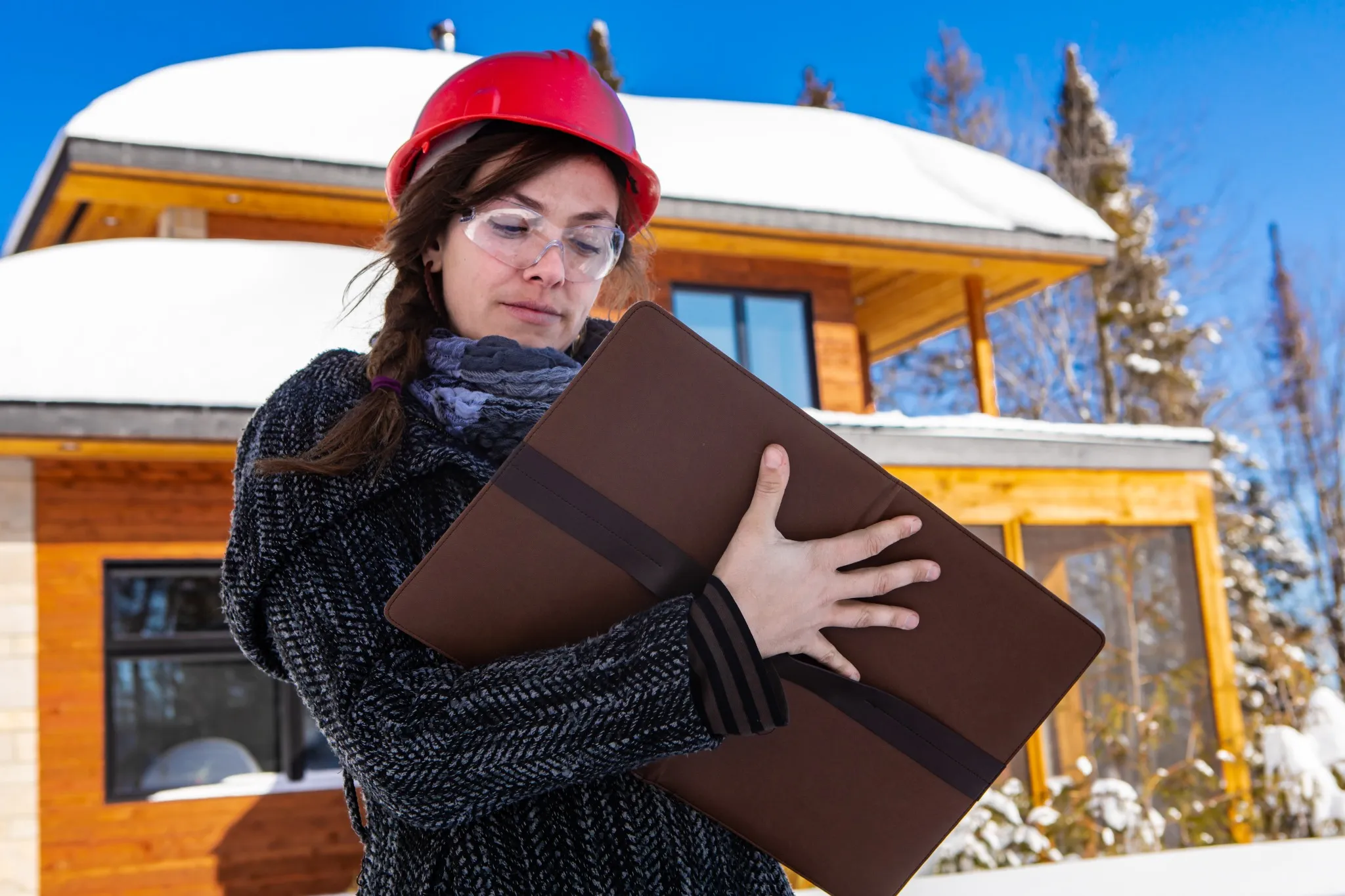 low angle view of a female civil engineer standing outdoor holding a clipboard and taking notes, the snow on wooden modern building in the background.