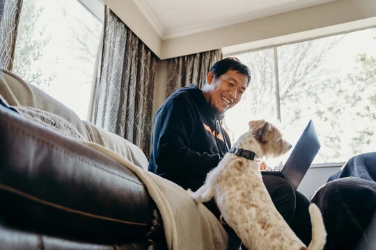 Smiling Asian man on couch with dog and laptop