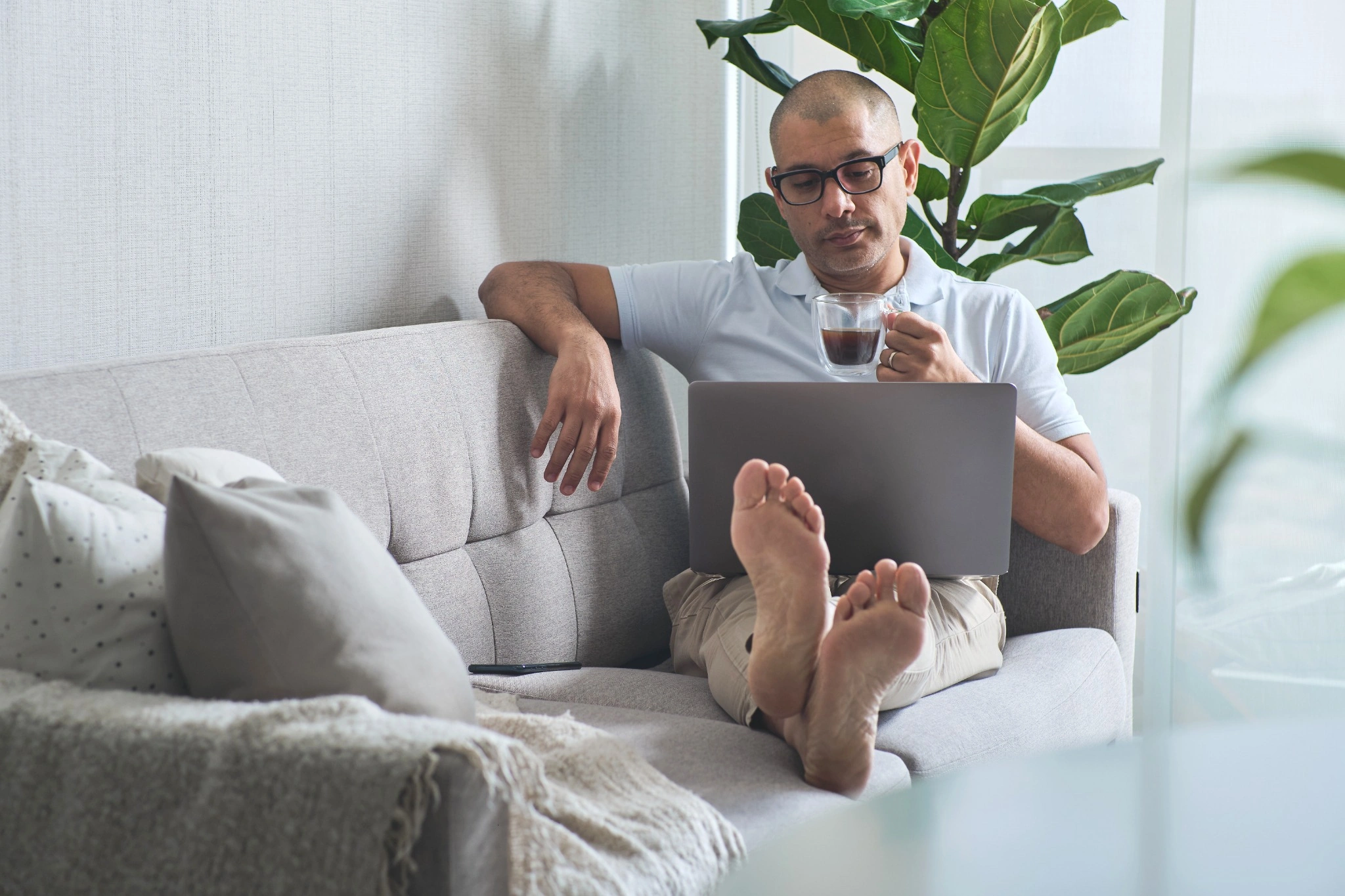Mature freelancer working from home, enjoying coffee and using laptop on sofa.