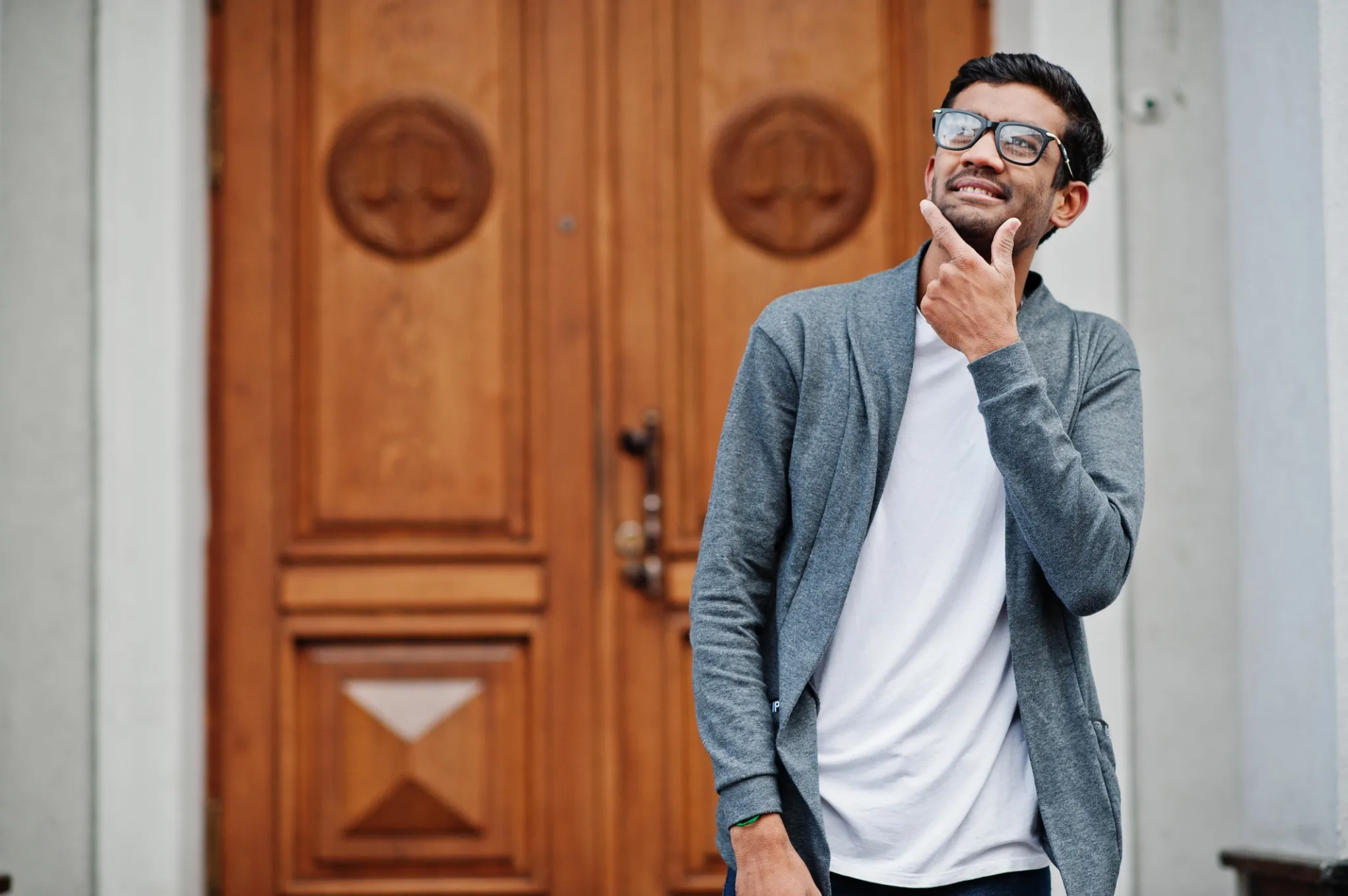 Stylish indian man at glasses wear casual posed outdoor against door of building, show that he thinking.