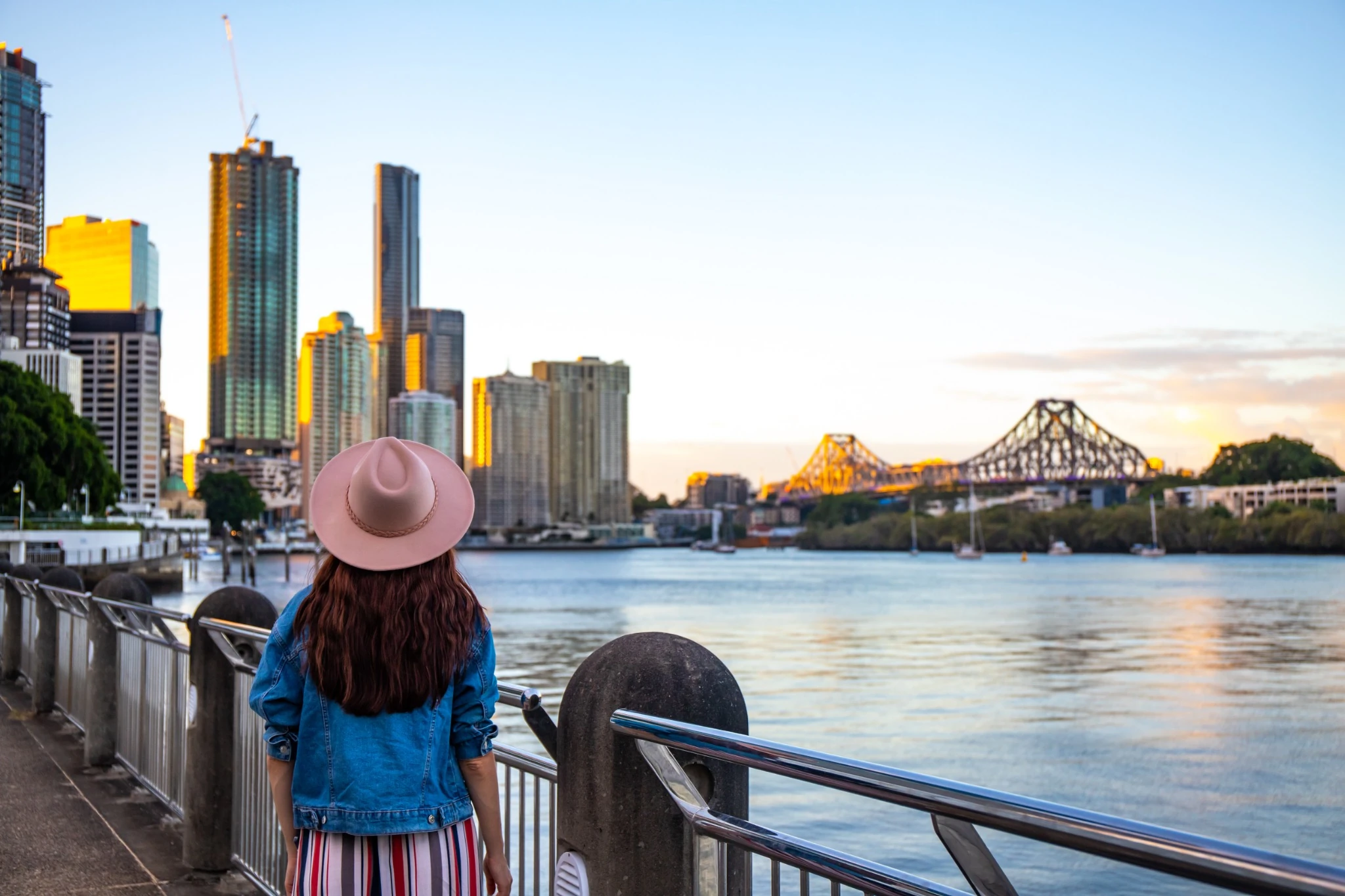 beautiful long hair woman in a hat watching the sunset over brisbane city; city reach boardwalk with amazing view of large skyscrapers by brisbane river, australia, holidays in brisbane.