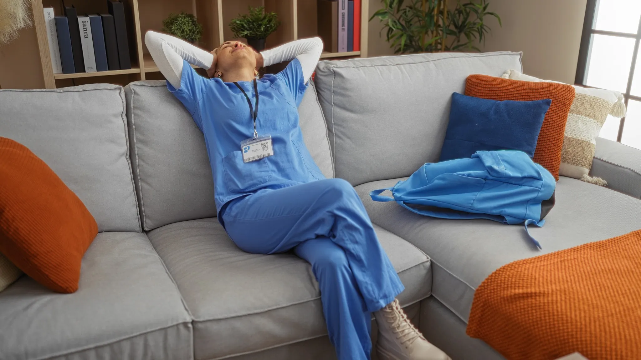 Woman relaxing on a sofa at home wearing blue scrubs with a backpack beside her in a cozy living room setting with cushions and bookshelves
