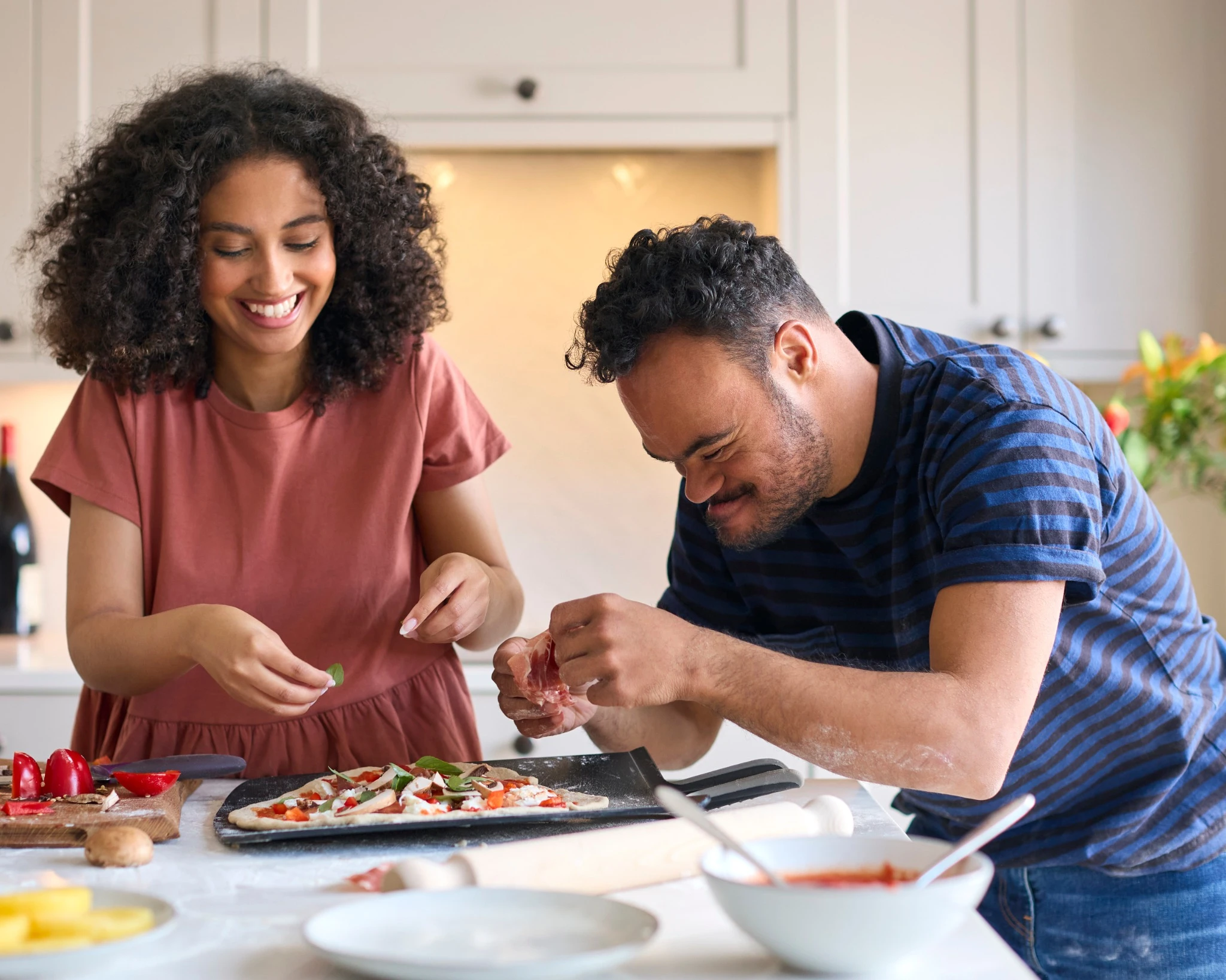Couple At Home With Man With Down Syndrome And Woman Putting Toppings On Pizza In Kitchen Together.