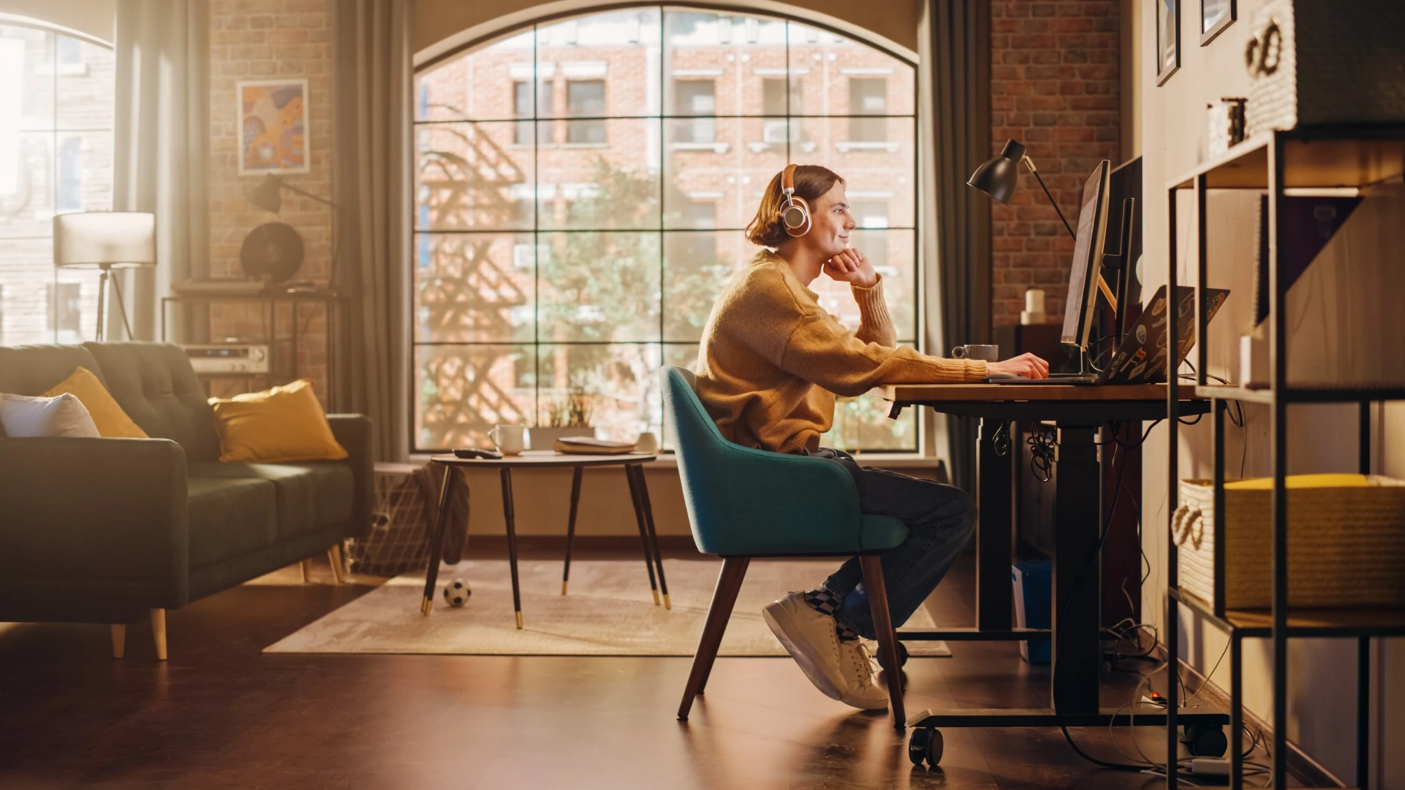 Young Handsome Man Working from Home on Desktop Computer in Sunny Stylish Loft Apartment. Creative Designer Wearing Cozy Yellow Sweater and Headphones. Urban City View from Big Window..