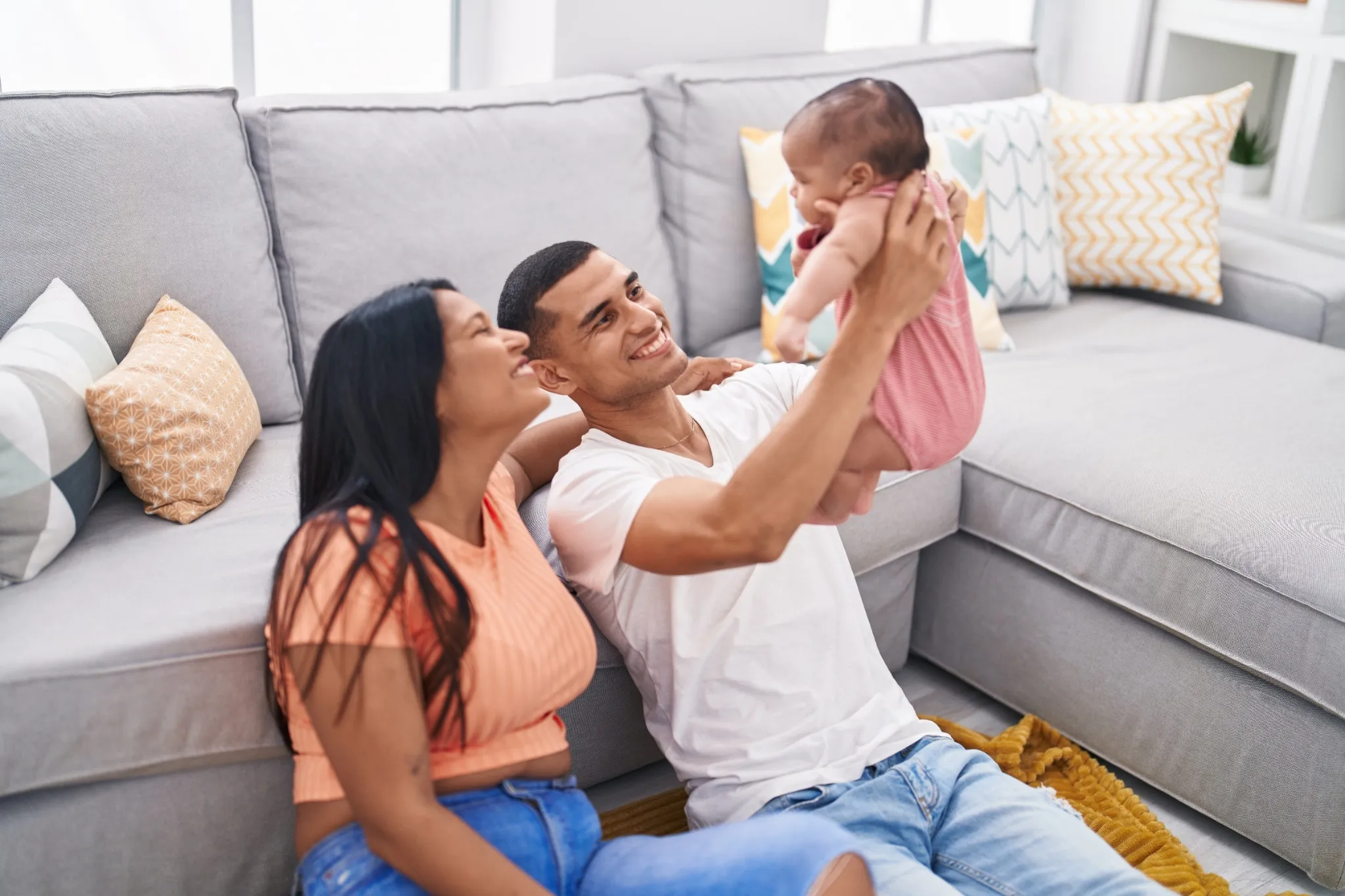 Hispanic family holding baby on air sitting on floor at home.