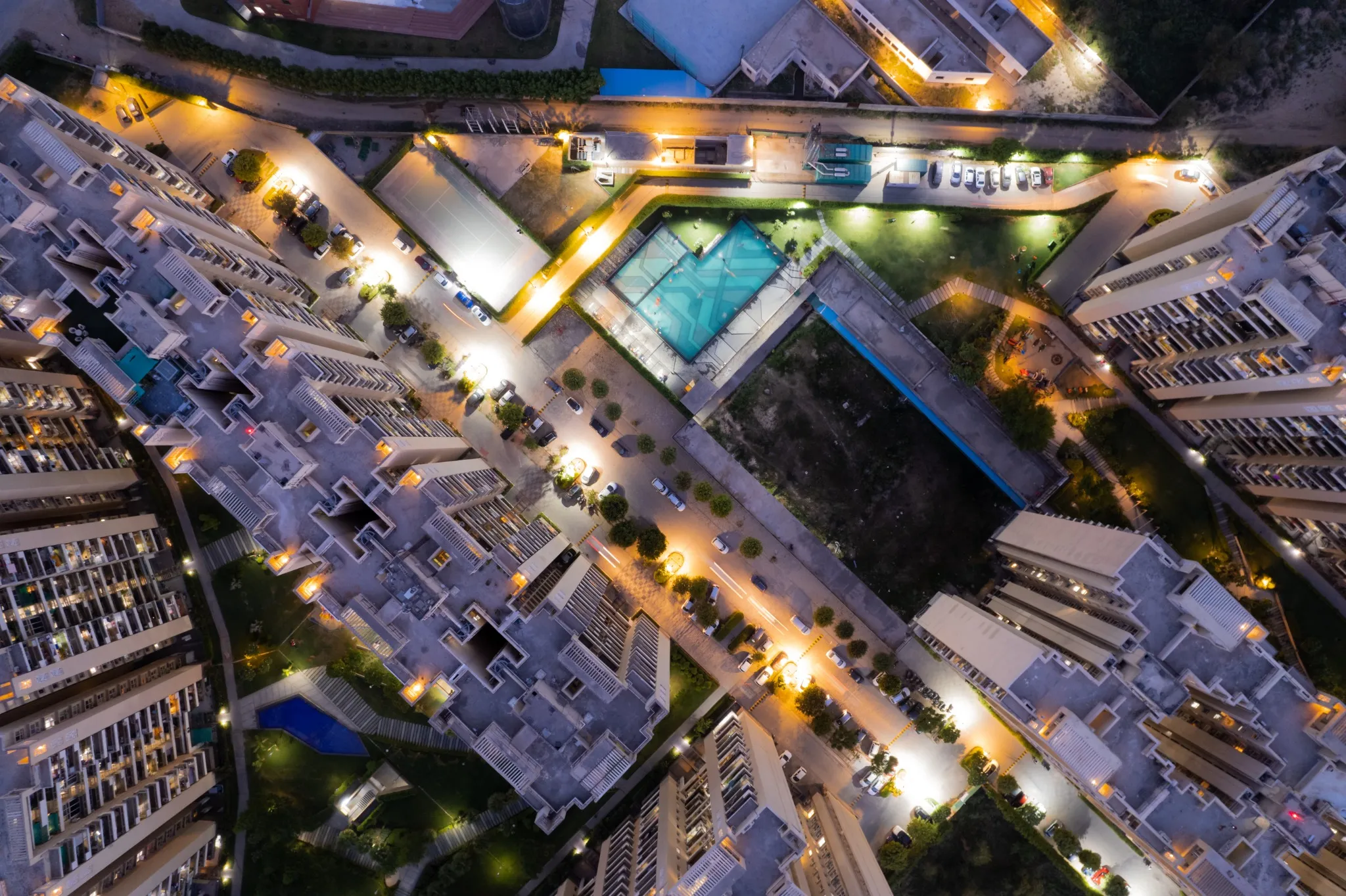 erial drone shot ooking down over skyscrapers of a premium apartment complex with parking swimming pools with lights switched on at dusk twilight in gurgaon haryana
