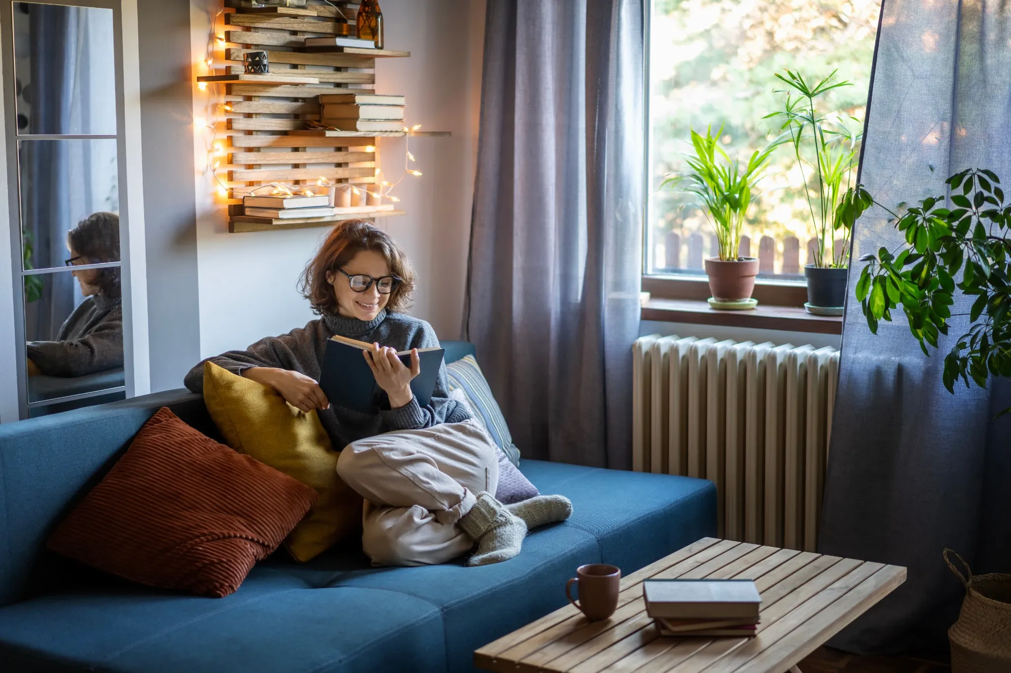 Young smiling cheerful woman in a warm sweater and eyeglasses reading a book while sitting on the couch in the room