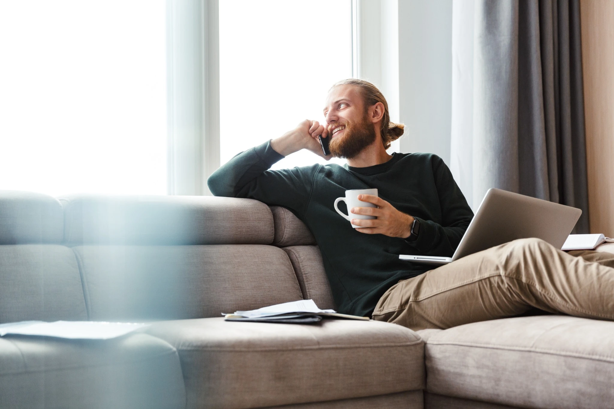 Man comfortable sitting on the couch talking on the phone with laptop and coffee.