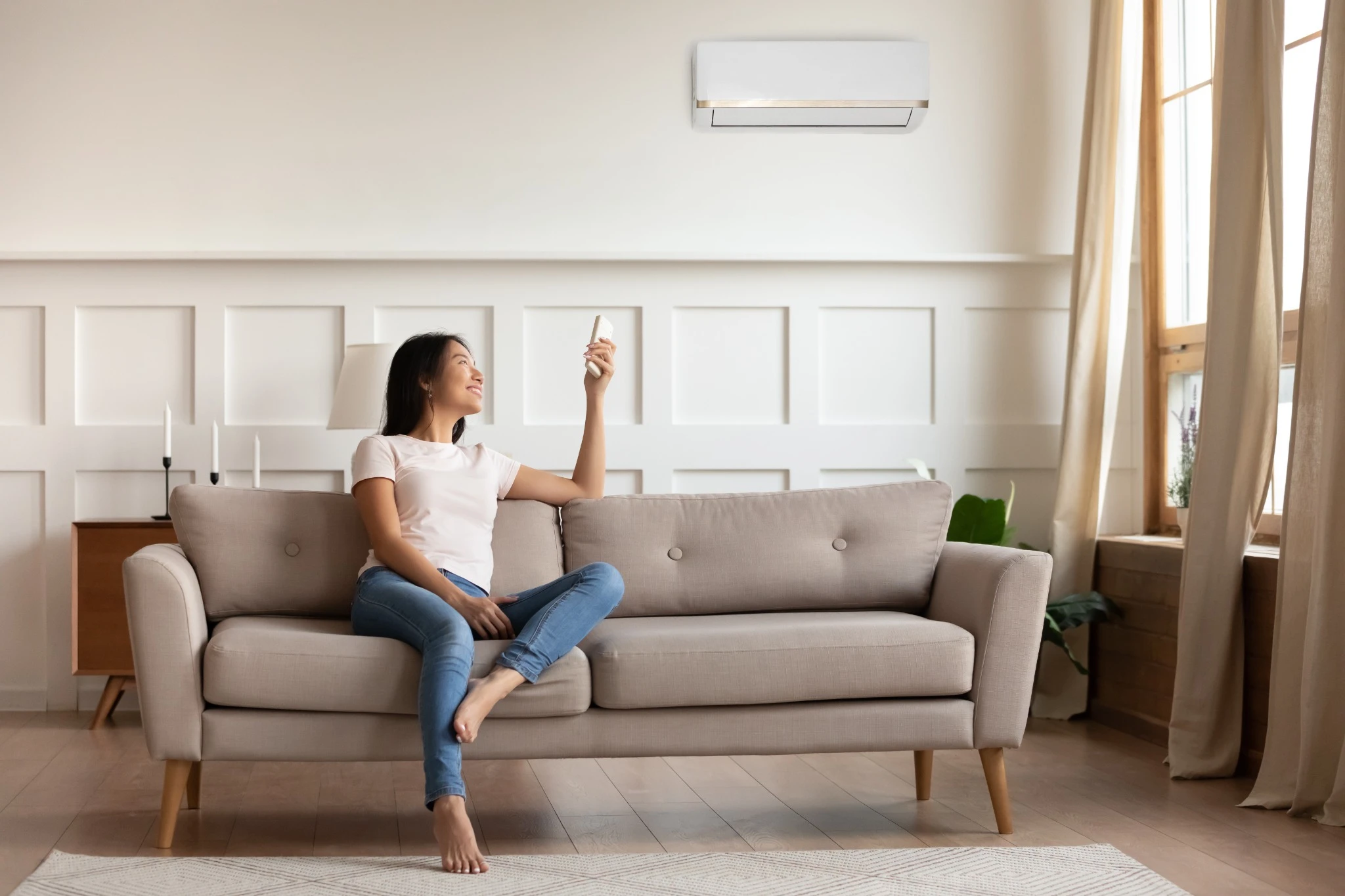 Young asian woman switching on air conditioner while resting seated on couch in modern interior living room, holds remote climate control cooler system manage temperature at contemporary home concept.